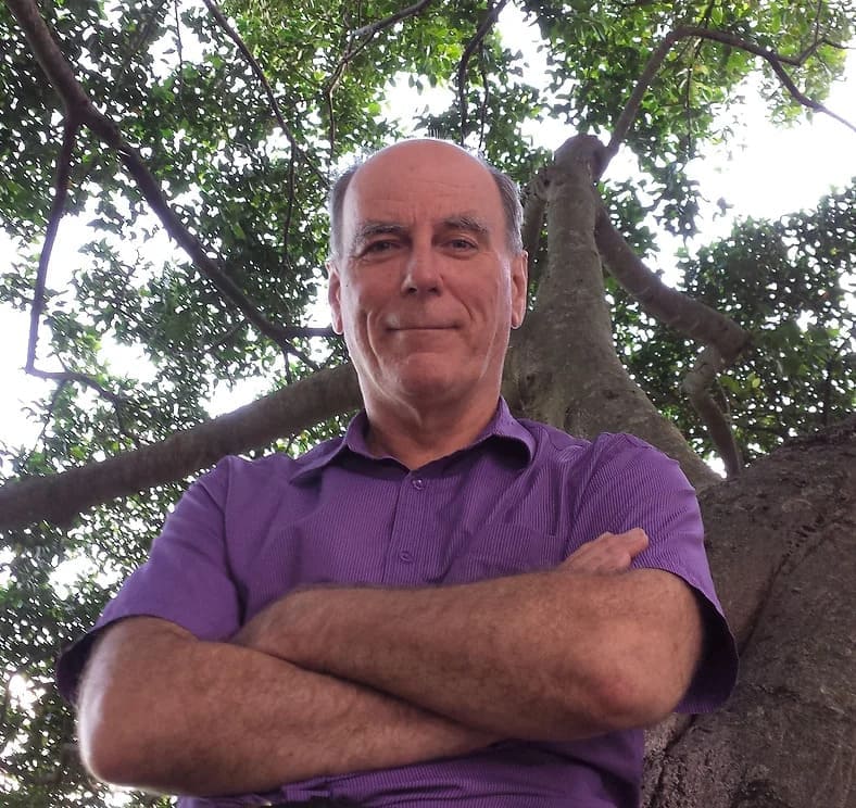 A man in a purple shirt sitting in front of a tree.